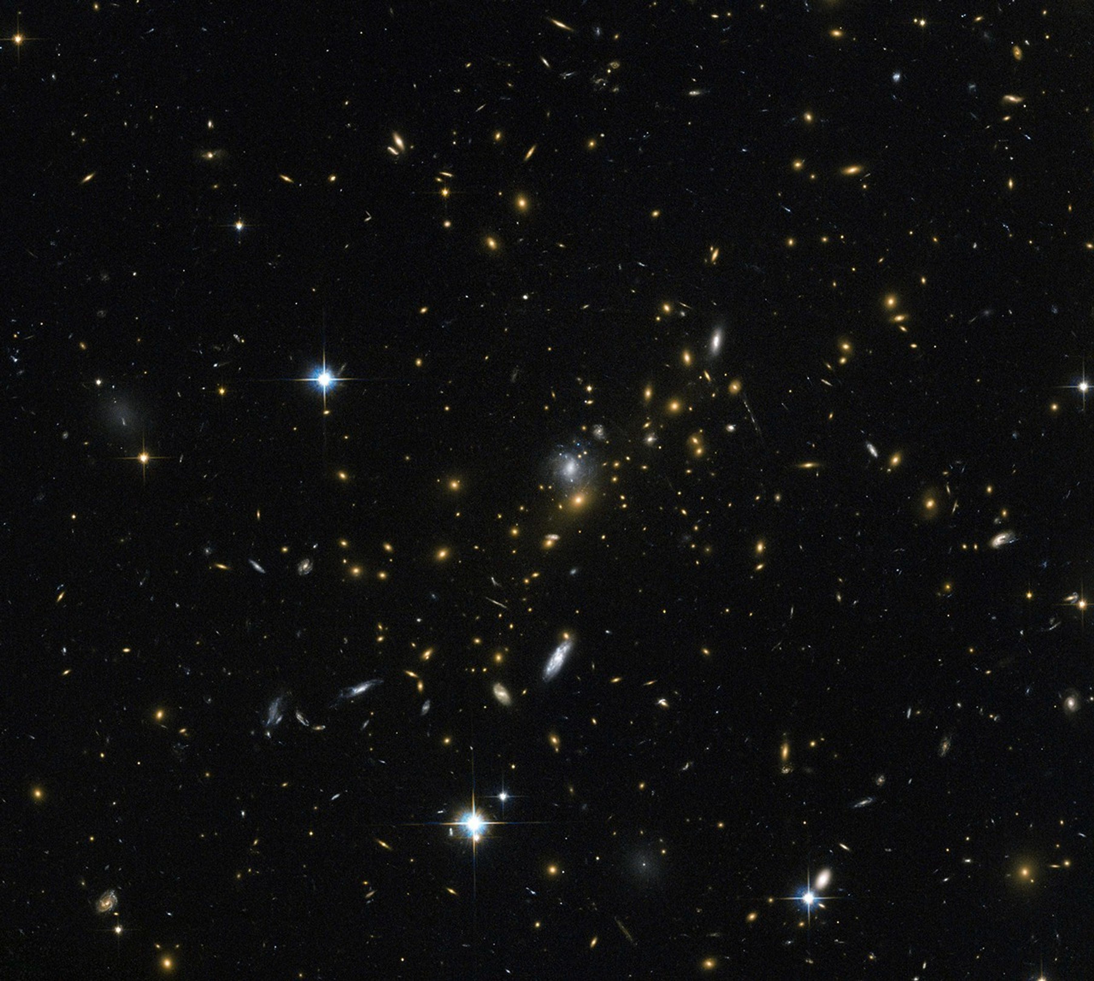 A field of galaxies and with faint curved arcs. Each galaxy appears as a glob of light, most of them reddish-orange. Bright, blue-white, twinkling foreground stars are scattered in a few places in the image.
